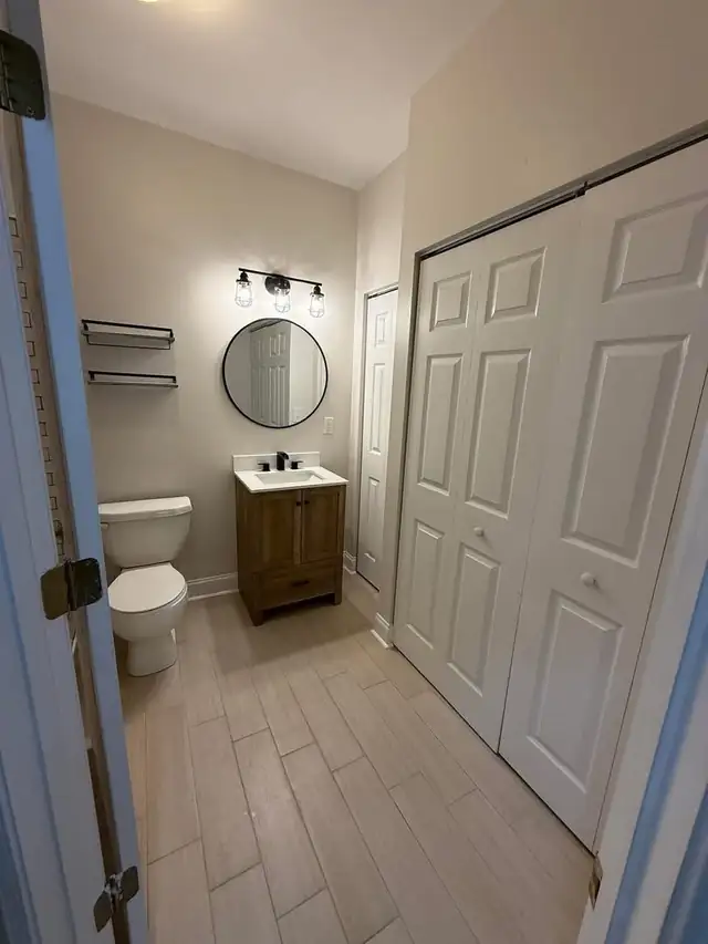 Remodeled small bathroom with wood vanity, round black mirror, black light fixture and wood-look tile floor